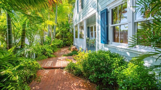 A white building with blue shutters, surrounded by lush greenery and a brick pathway. - Passover Cottage | Private Pool & Ideal Key West Location (Key West)