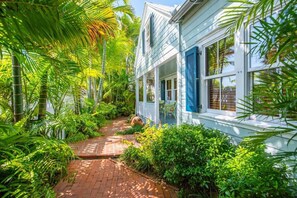 A white building with blue shutters, surrounded by lush greenery and a brick pathway. - Passover Cottage | Private Pool & Ideal Key West Location (Key West)