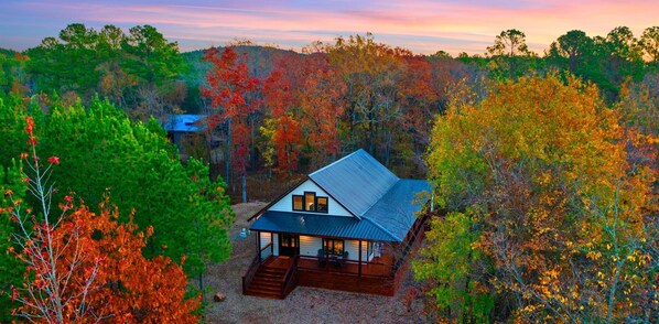 A cabin with a sloped roof, surrounded by trees with autumn foliage, and a clear sky. - Games | Playset | Hot Tub | Wraparound Porch (Broken Bow)