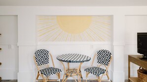 A room with a white wall, a round table, three patterned chairs, a large abstract painting, and a television on a wooden stand.