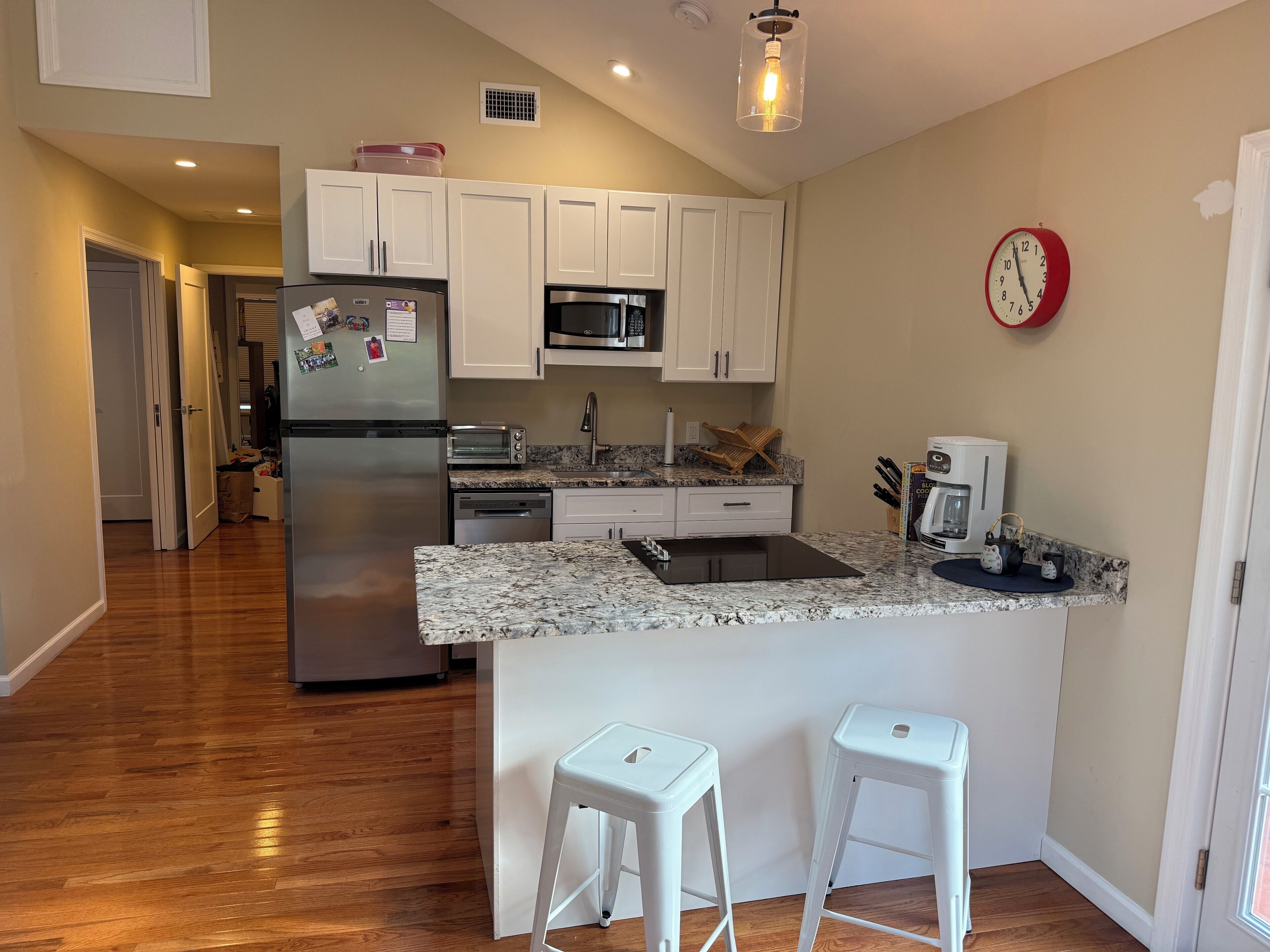 A modern kitchen with white cabinets, a stainless steel refrigerator, a microwave, a toaster, and a coffee maker. There are two white bar stools and a clock on the wall.
