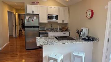A modern kitchen with white cabinets, a stainless steel refrigerator, a microwave, a toaster, and a coffee maker. There are two white bar stools and a clock on the wall.