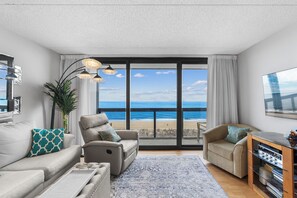 A modern living room with a large window offering a beach view, a sofa, a chair, and a TV stand.