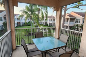 Un balcón con una mesa y sillas de vidrio, con vistas a una zona residencial con casas y palmeras.