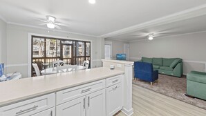 Une cuisine moderne avec des armoires blanches, un grand îlot central et un coin repas avec des chaises blanches. Le salon est agrémenté d’un canapé vert et d’un fauteuil bleu.