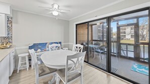 Une cuisine moderne avec des meubles blancs, une table à manger blanche accompagnée de quatre chaises, et une grande porte-fenêtre coulissante donnant sur un patio extérieur.