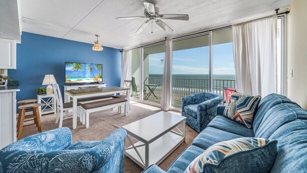 Un salon avec un canapé bleu, une table basse blanche, un téléviseur et un balcon offrant une vue sur l’océan.