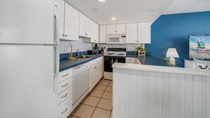 Une cuisine moderne avec des armoires blanches, un four à micro-ondes et une cuisinière. On y trouve un évier et un plan de travail avec une machine à café. La pièce a des murs bleus et une fenêtre donnant sur la mer.