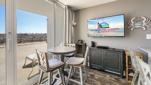 A spacious room with a large sliding glass door leading to a balcony, a round table with chairs, a television mounted on the wall, and a small cabinet.
