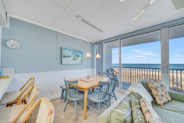 A room with a dining table, chairs, a sofa, and a view of the beach.
