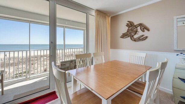 A room with a wooden table and white chairs, a large window with a view of the beach, and a wall-mounted decorative piece. - Sea Gate 205 by Seaside Vacations & Sales (Ocean City)