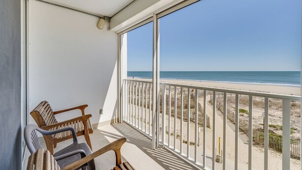 A balcony with a view of the beach, wooden chairs, and a white railing. - Sea Gate 205 by Seaside Vacations & Sales (Ocean City)