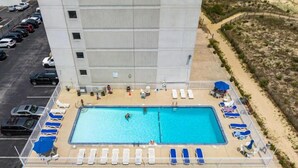 An aerial view of a hotel with a swimming pool surrounded by sun loungers, a parking lot, and a sandy area with umbrellas. - Stunning condo steps from the beach by Seaside Vacations & Sales (Ocean City)