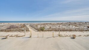 Vista de la playa con un camino de hormigón que lleva al agua, rodeado de dunas de arena y una valla de madera.