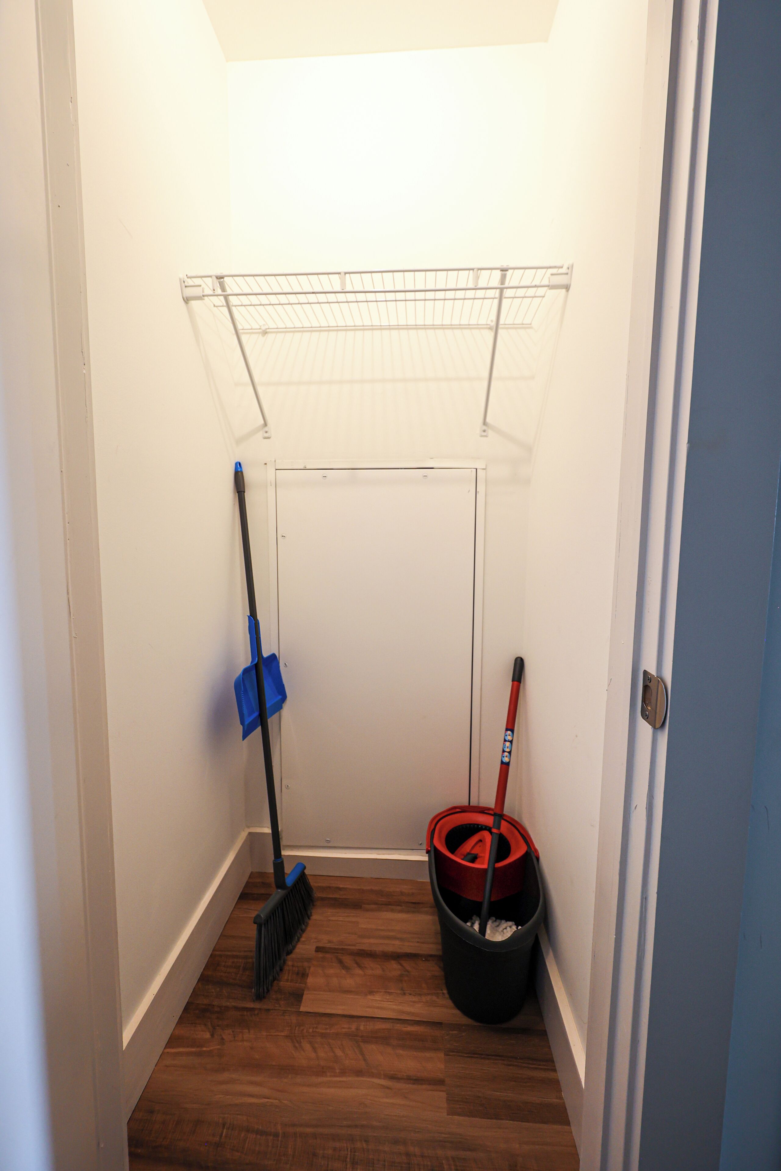 A narrow hallway with a wooden floor, a metal drying rack, a blue dustpan, a red-handled broom, and a black trash bin.