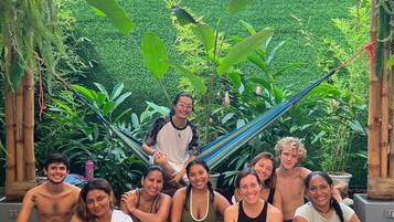 A group of people sitting on exercise mats outdoors, surrounded by greenery and a bamboo structure.