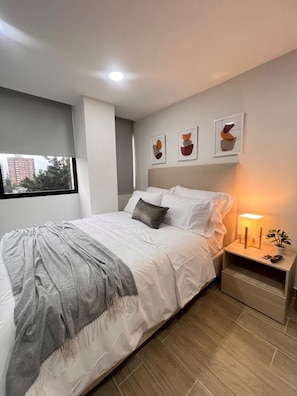 A neatly made bed with a gray blanket, a bedside lamp, and a small plant on a wooden nightstand. - 81 Living (Medellín)