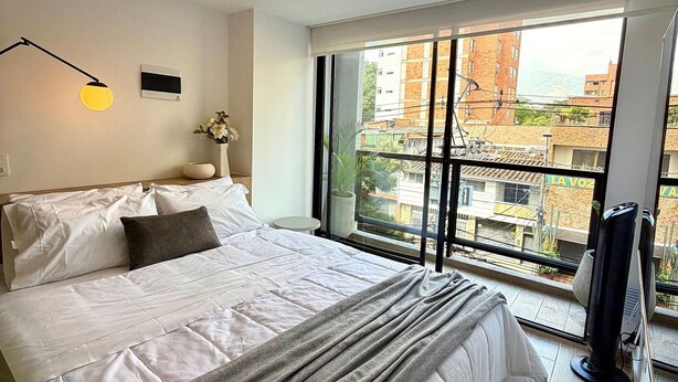 Een slaapkamer met een groot bed, een nachtkastje, een lamp en uitzicht op gebouwen door een raam.