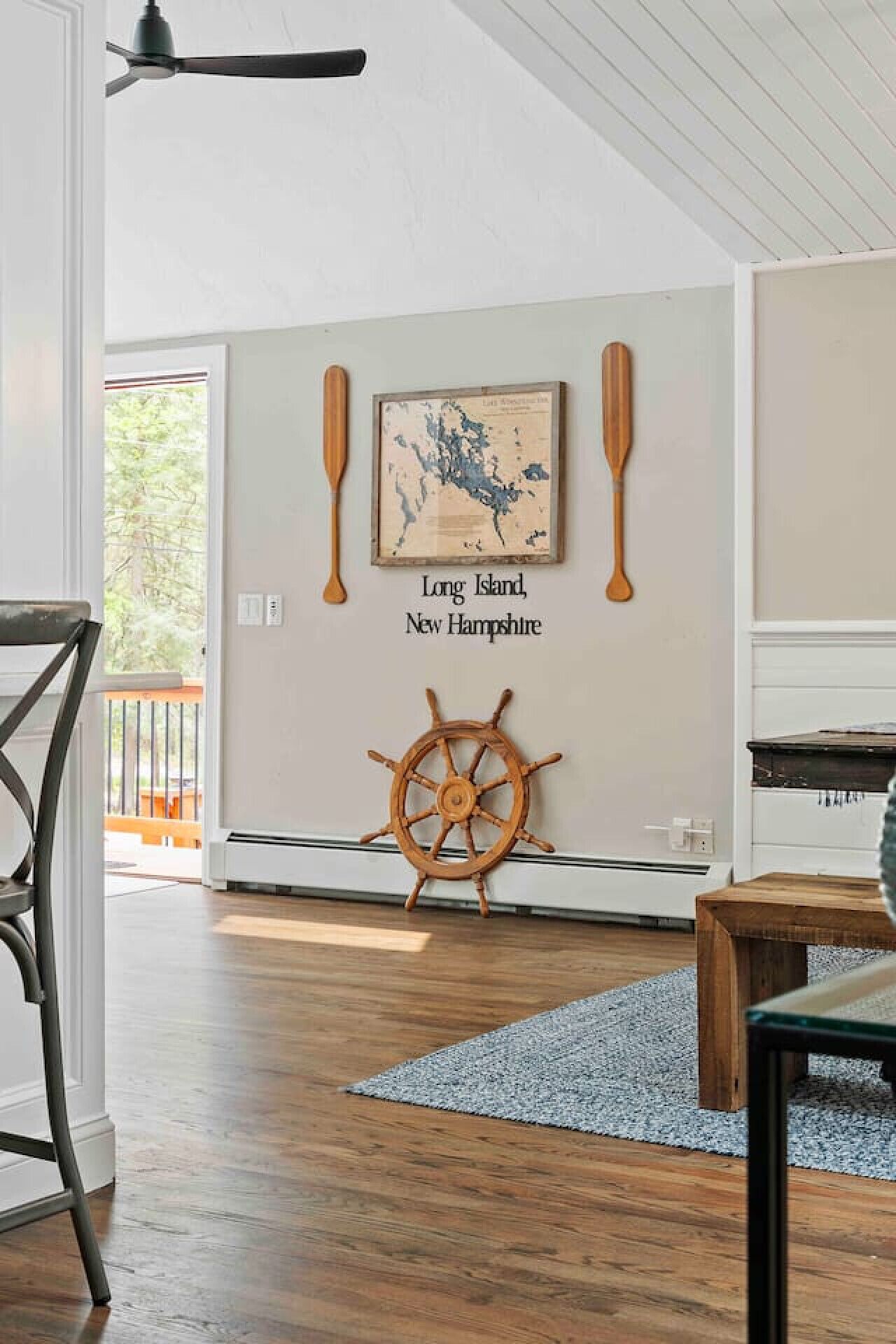 Une pièce avec un plancher en bois, une carte encadrée de Long Island et du New Hampshire, une pagaie en bois et une roue de navire.