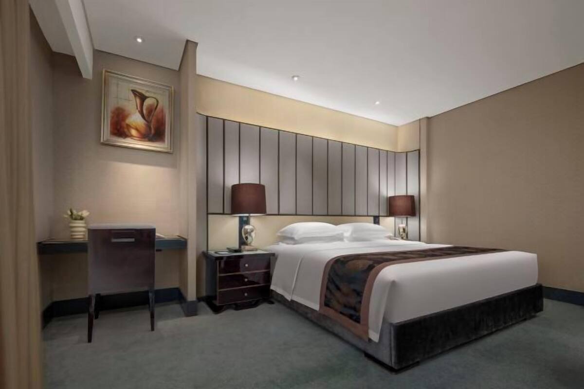A hotel room with a bed, bedside lamps, a desk, and a painting on the wall.