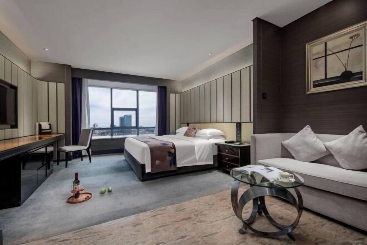 A modern hotel room with a bed, a desk, a sofa, a glass coffee table, and a large window with a city view.