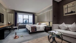 Habitación de hotel moderna con cama, escritorio, sofá, mesa de centro de vidrio y ventana grande con vista a la ciudad.