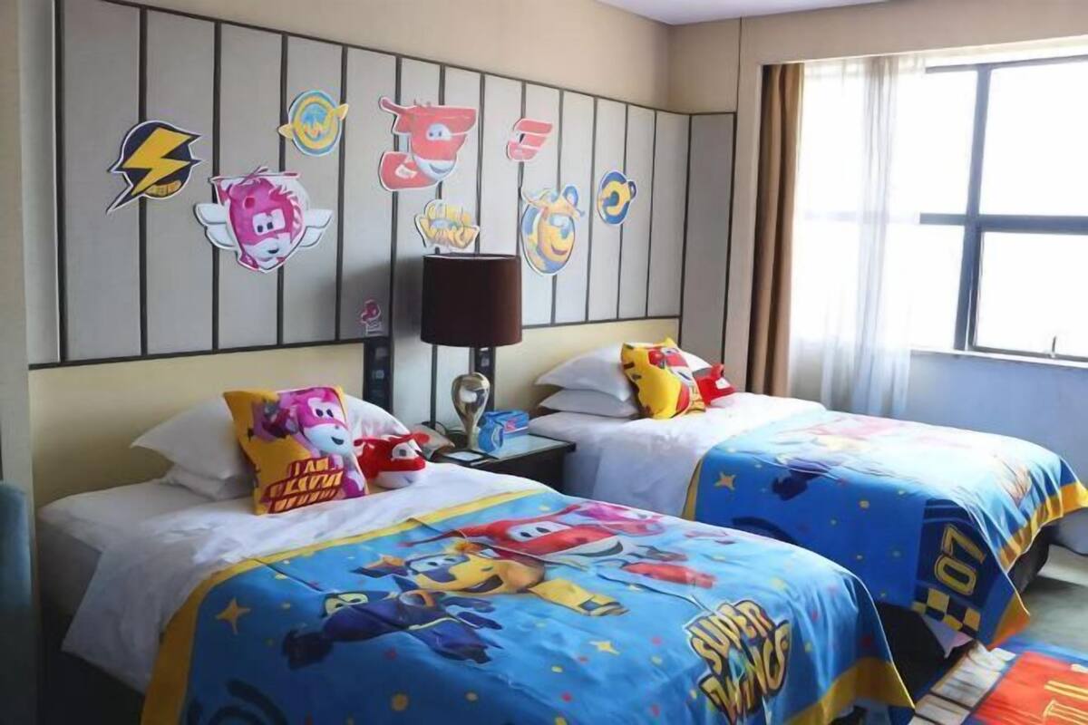 A hotel room with two beds, each with a cartoon-themed bedspread, a wall with cartoon character decorations, a nightstand with a lamp, and a window with curtains.
