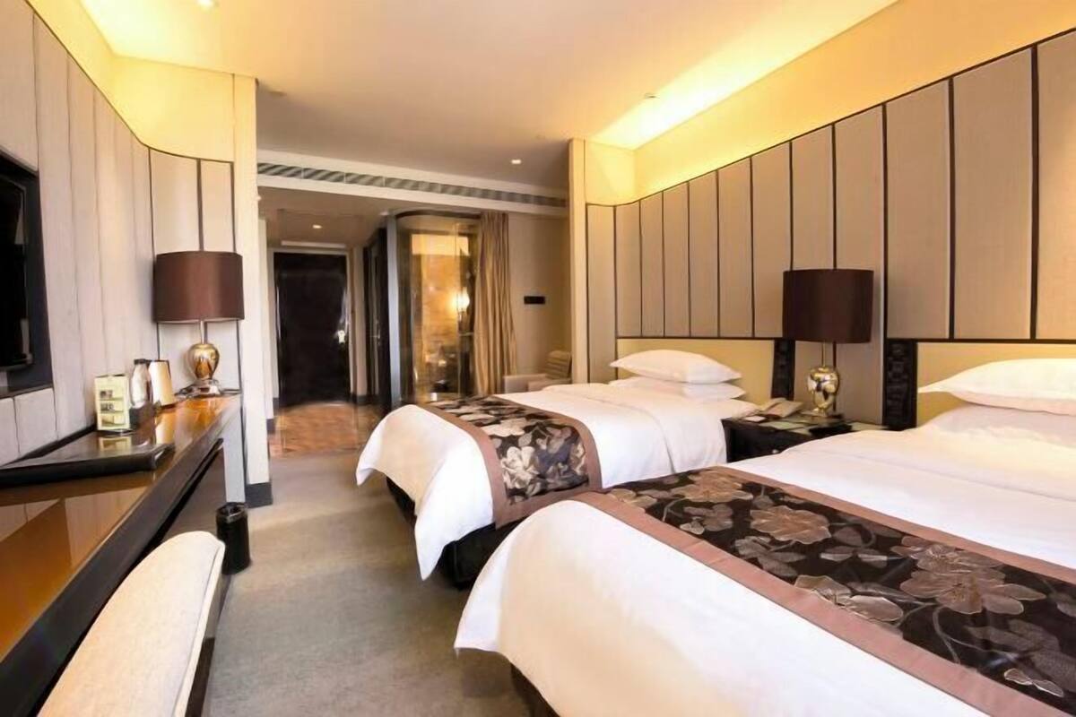 A hotel room with three beds, a desk with a lamp, a mirror, and a door leading to another room.