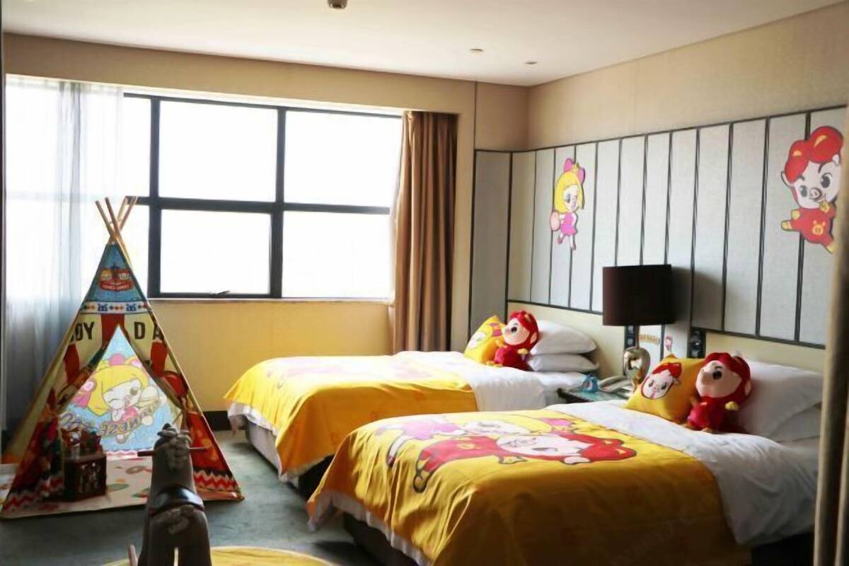 A hotel room with two beds, each with yellow bedding and cartoon character pillows. There is a window with curtains, a wall-mounted TV, and a small table with a lamp.
