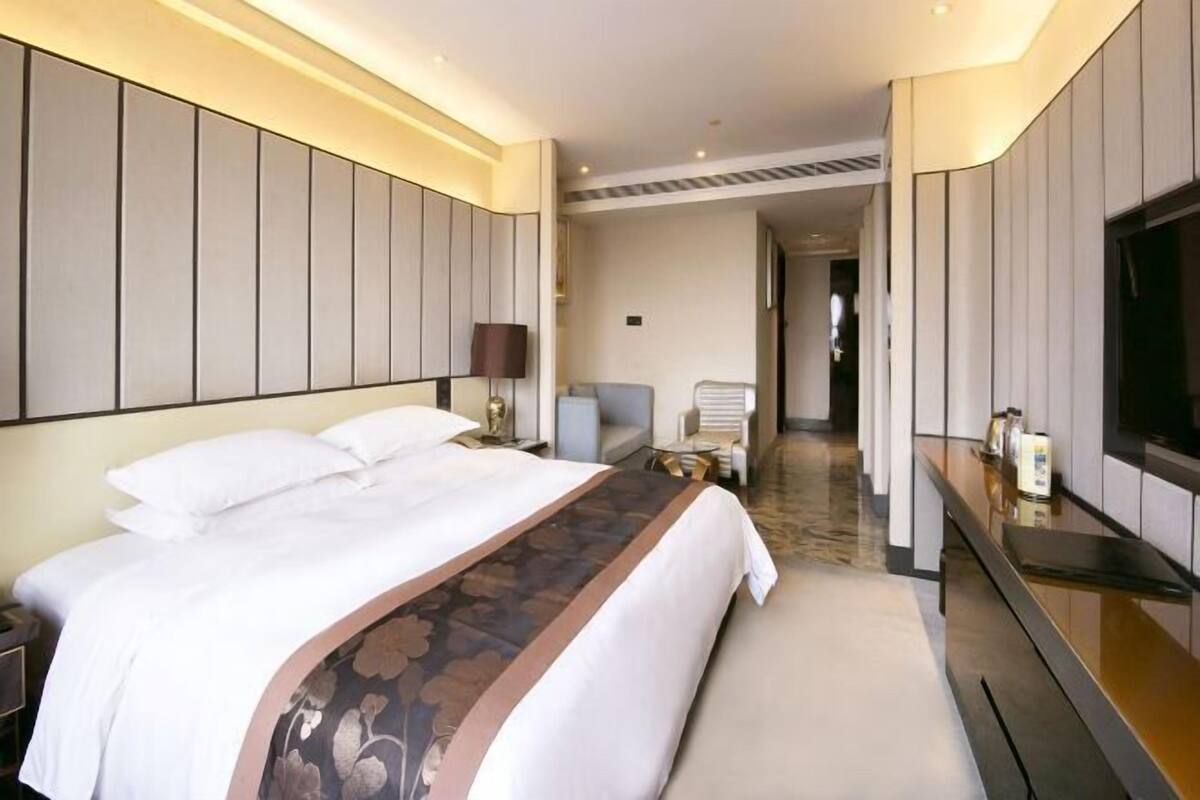 A hotel room with a large bed, a desk with a flat-screen TV, and a seating area with a chair and a small table.