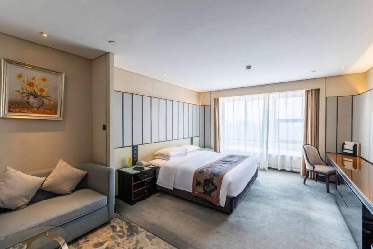 A modern hotel room with a large bed, a sofa, a desk, and a large window with curtains.