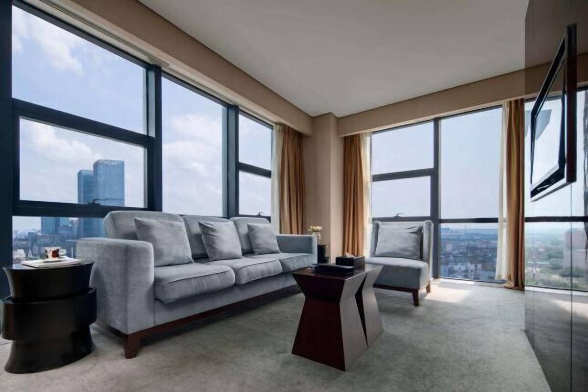 A modern living room with a grey sofa, a coffee table, and a chair. Large windows offer a city view.
