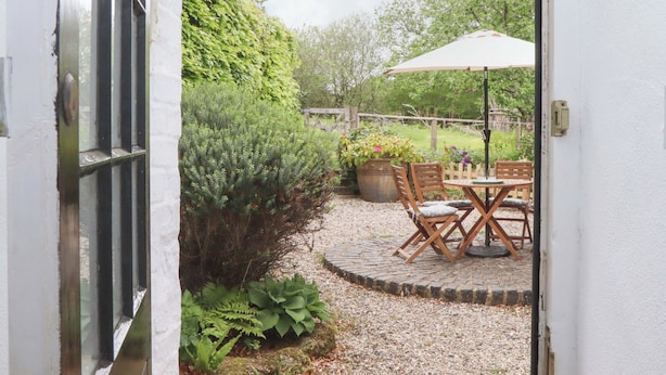 An open door leading to a patio with a table and chairs, surrounded by greenery and a wooden fence. - 2 New Mill Cottages (Newton Abbot)