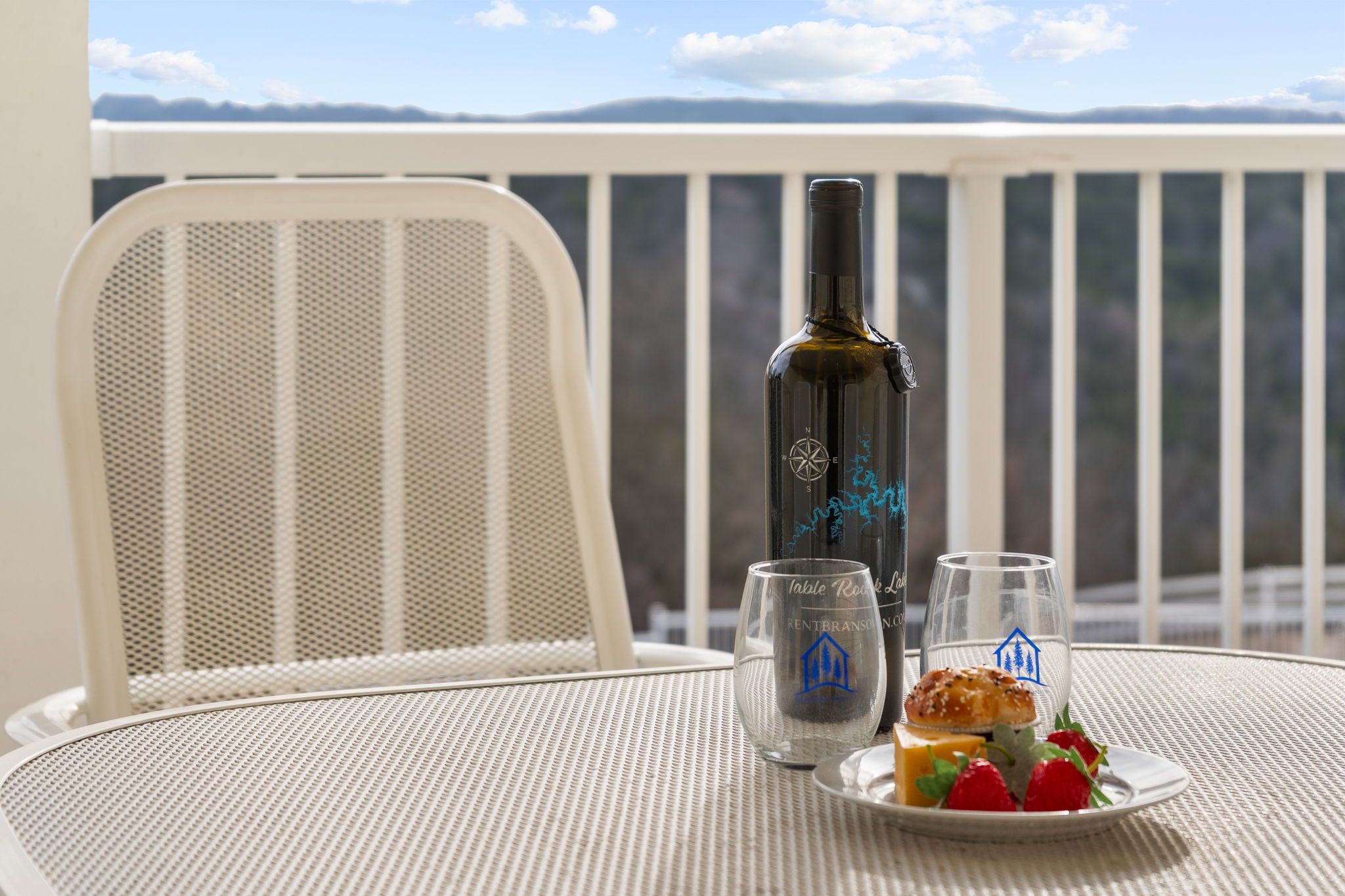 A table with a bottle, glasses, and a plate with food on a balcony overlooking a mountainous landscape.