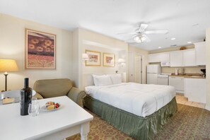 Quarto de hotel com cama, mesa de jantar, garrafa e copos, cozinha compacta e ventilador de teto.