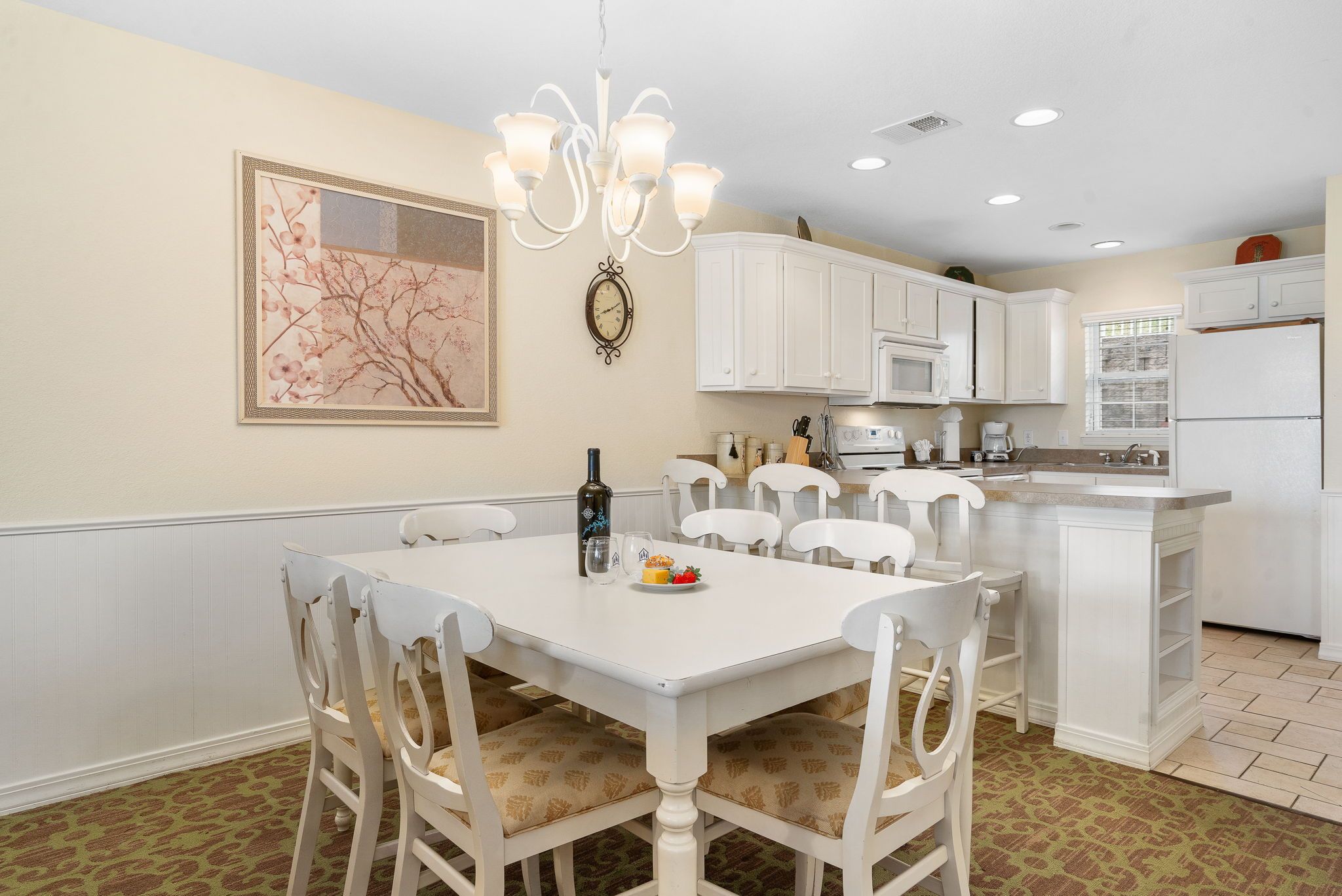 A kitchen with white cabinets, a white dining table, and chairs. There is a clock on the wall and a chandelier above the table.