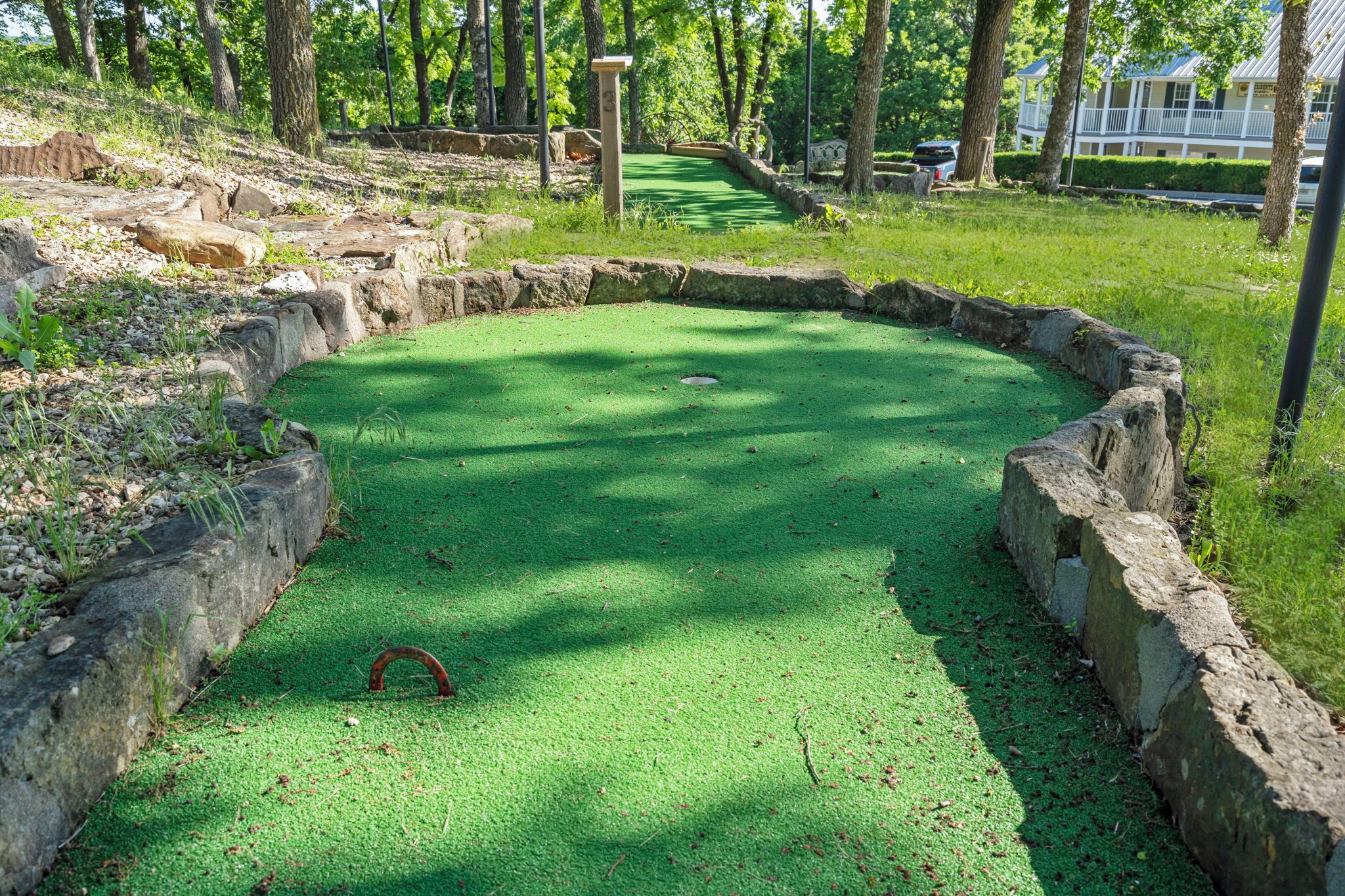 A miniature golf course with a green putting surface, surrounded by stone walls and trees.
