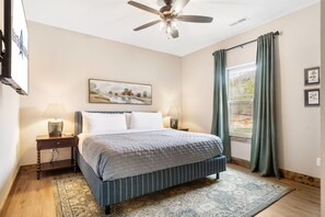 Une chambre à coucher avec un lit, un ventilateur de plafond, une fenêtre avec des rideaux, un tableau au mur et une table de chevet avec une lampe.