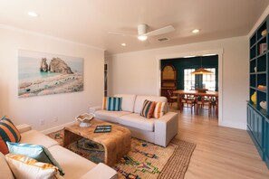 Un salon avec un canapé blanc, une table basse en bois et un tapis coloré. Un tableau représentant une scène de plage est accroché au mur.