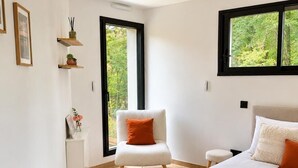 Ein Schlafzimmer mit einem großen Bett, einem Sessel, einem Nachttisch und einem Fenster mit Blick auf Grünflächen.