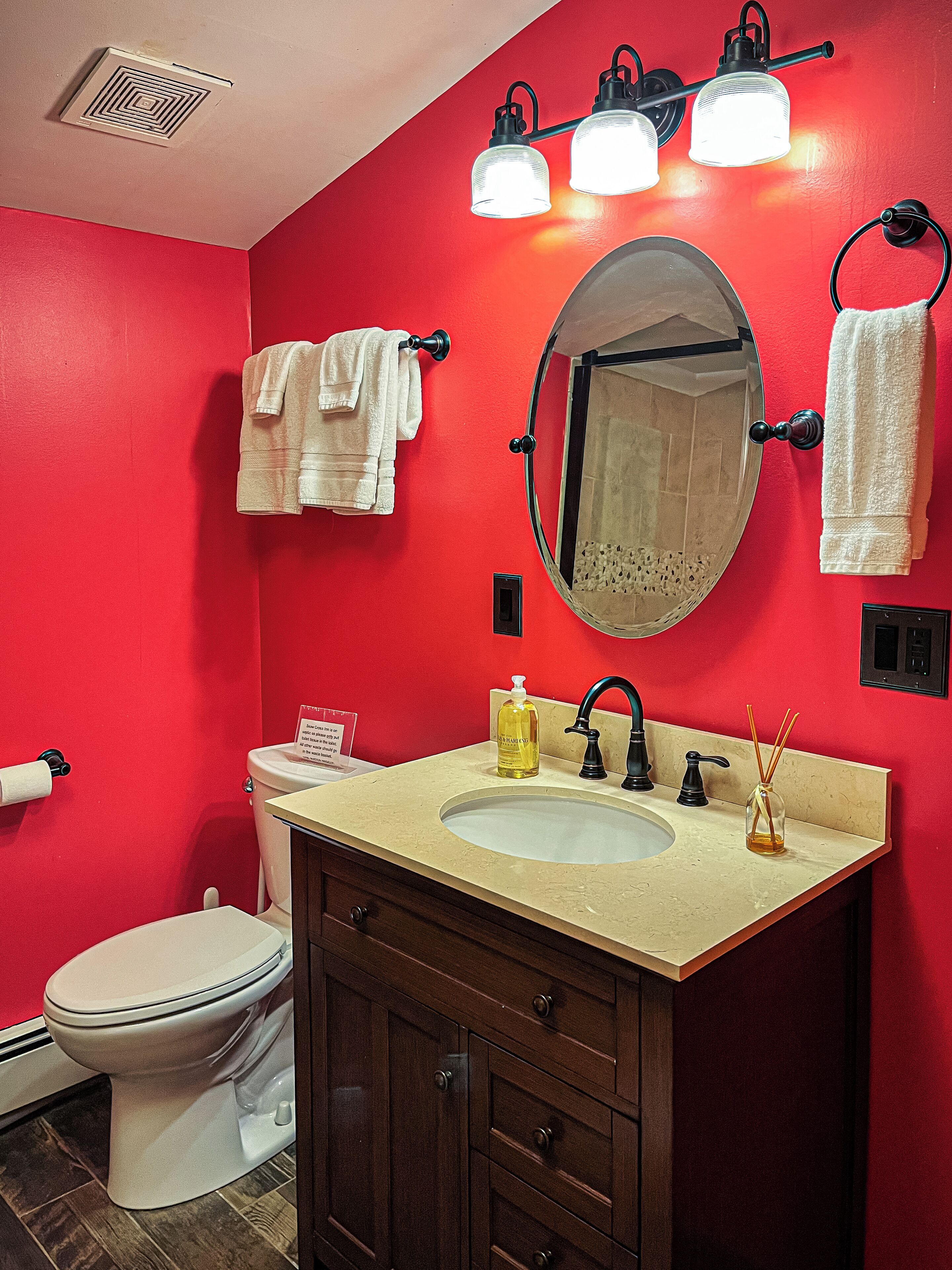Ein Badezimmer mit roter Wand, einer weißen Toilette, einem beige gefärbten Waschbecken mit Spiegel darüber und zwei weißen Handtüchern, die an Handtuchhaken hängen.