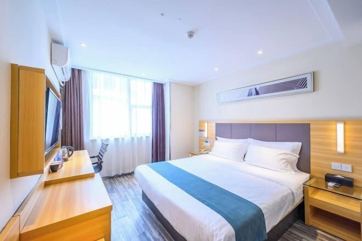 A hotel room with a bed, bedside tables, a desk, a chair, a TV, and a window with curtains.