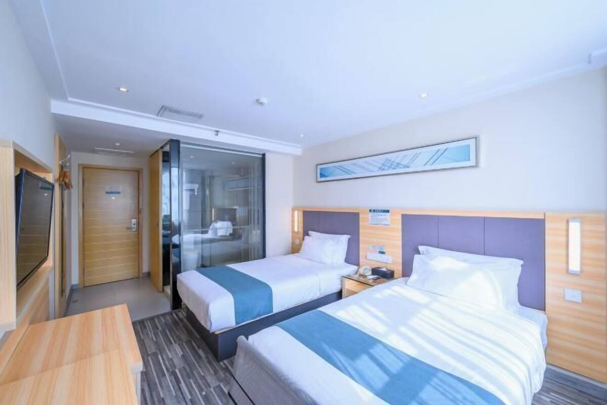 A hotel room with two beds, a desk, a glass partition, and a framed picture on the wall.
