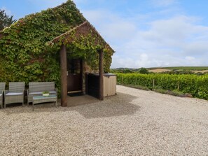 A rustic building with a thatched roof, covered in ivy, and a gravel driveway leading to a seating area with wicker furniture. - Babes Cottage (Truro)
