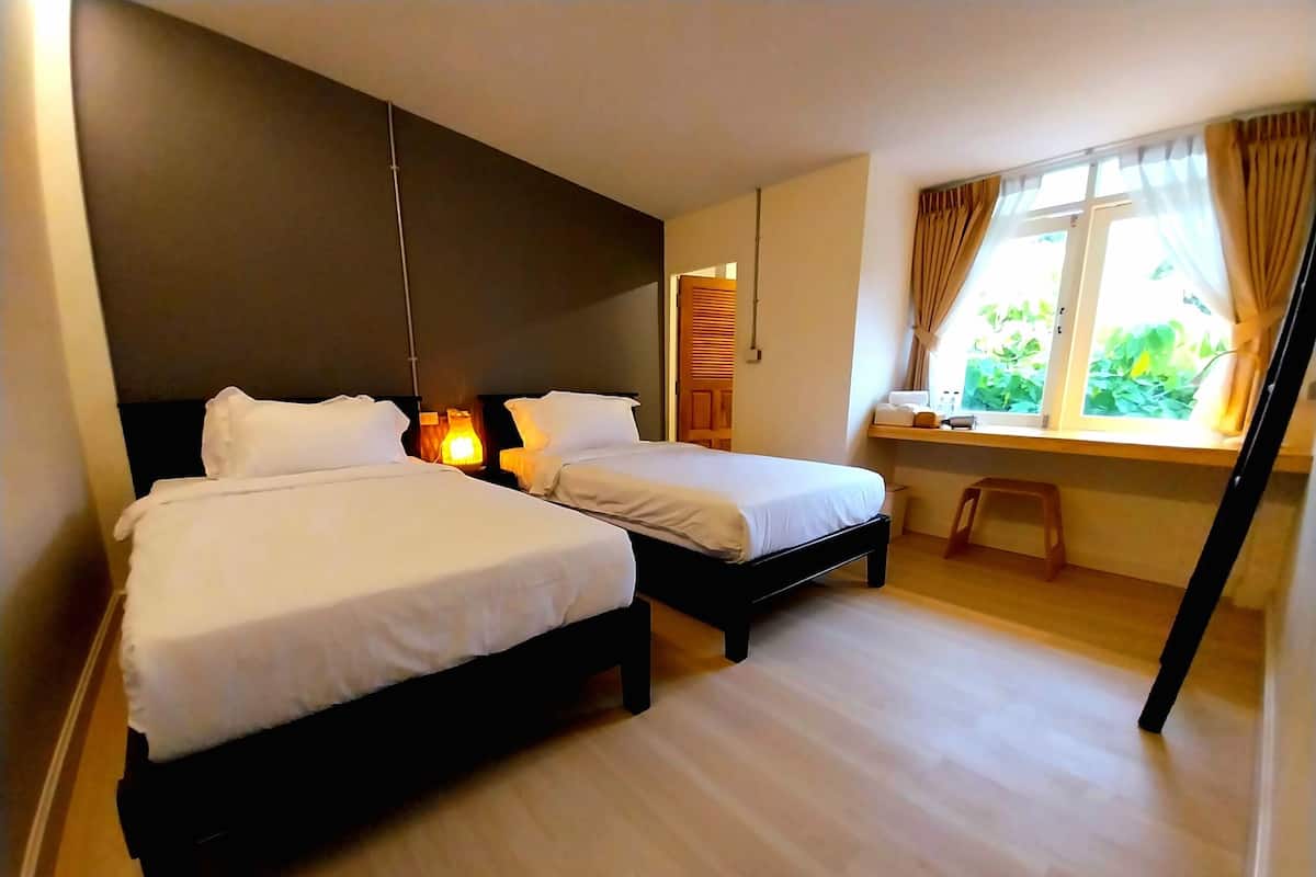 A hotel room with two beds, a desk, a window with curtains, and a small stool.