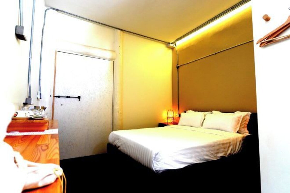 A small, compact hotel room with a single bed, a bedside table, and a nightstand. There is a door with a handle and a light switch on the wall.