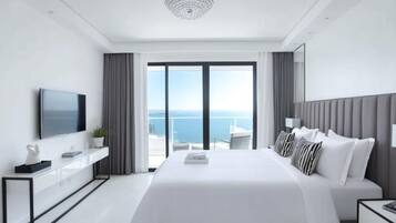 A modern hotel room with a large bed, a flat-screen TV, a bedside table, and a view of the sea through large windows.