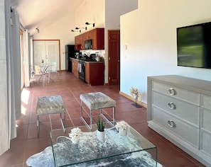 Una cuina moderna amb armaris de fusta, una zona de menjador amb taula de vidre i un saló amb televisió i sofà.