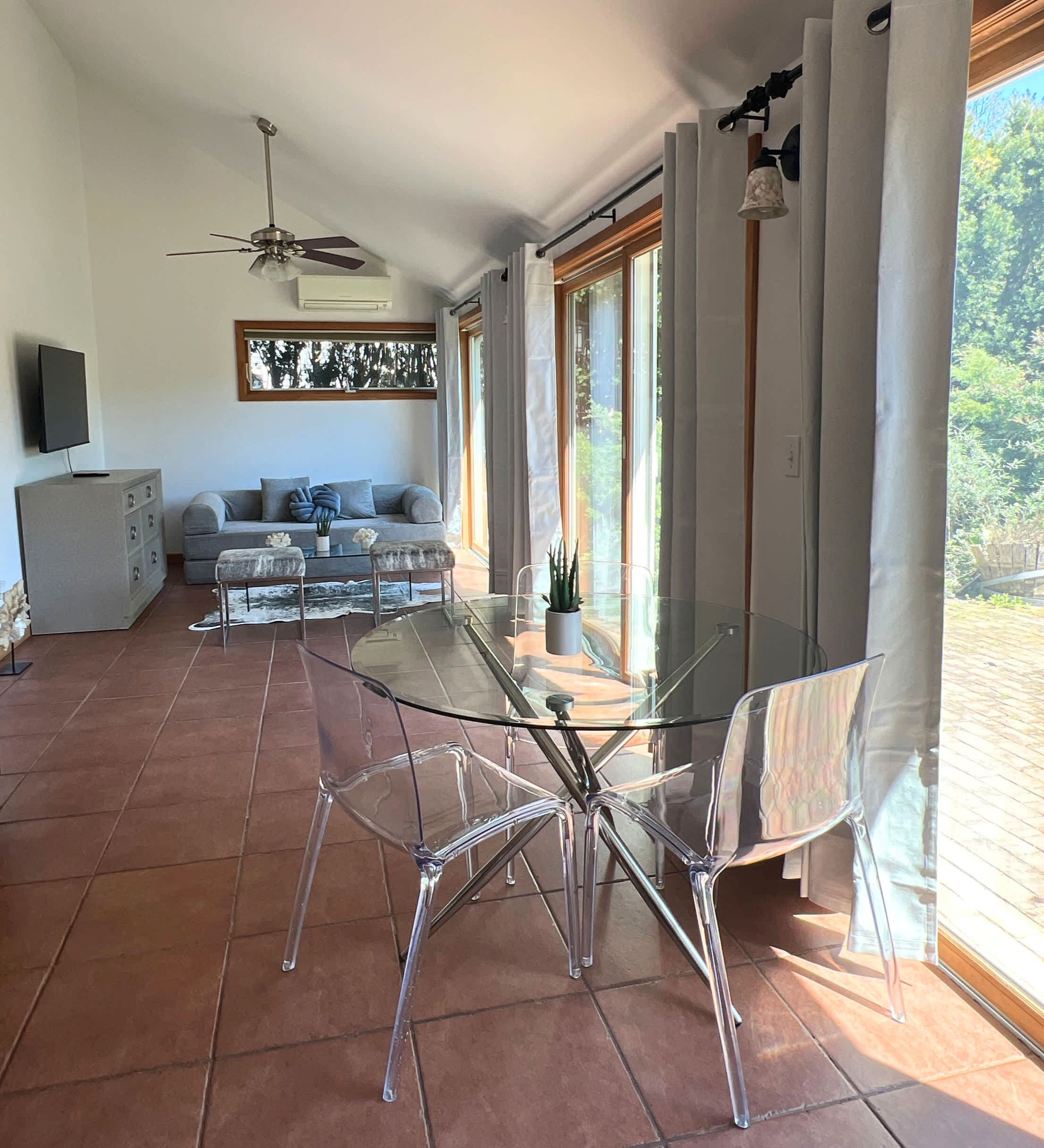 Una sala moderna con una mesa de vidrio, sillas transparentes, un sof á gris y un ventilador de techo.