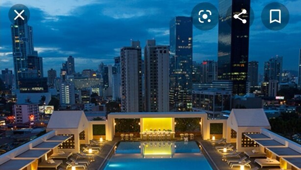 A rooftop pool area with lounge chairs and tables, a bar, and a city skyline view.